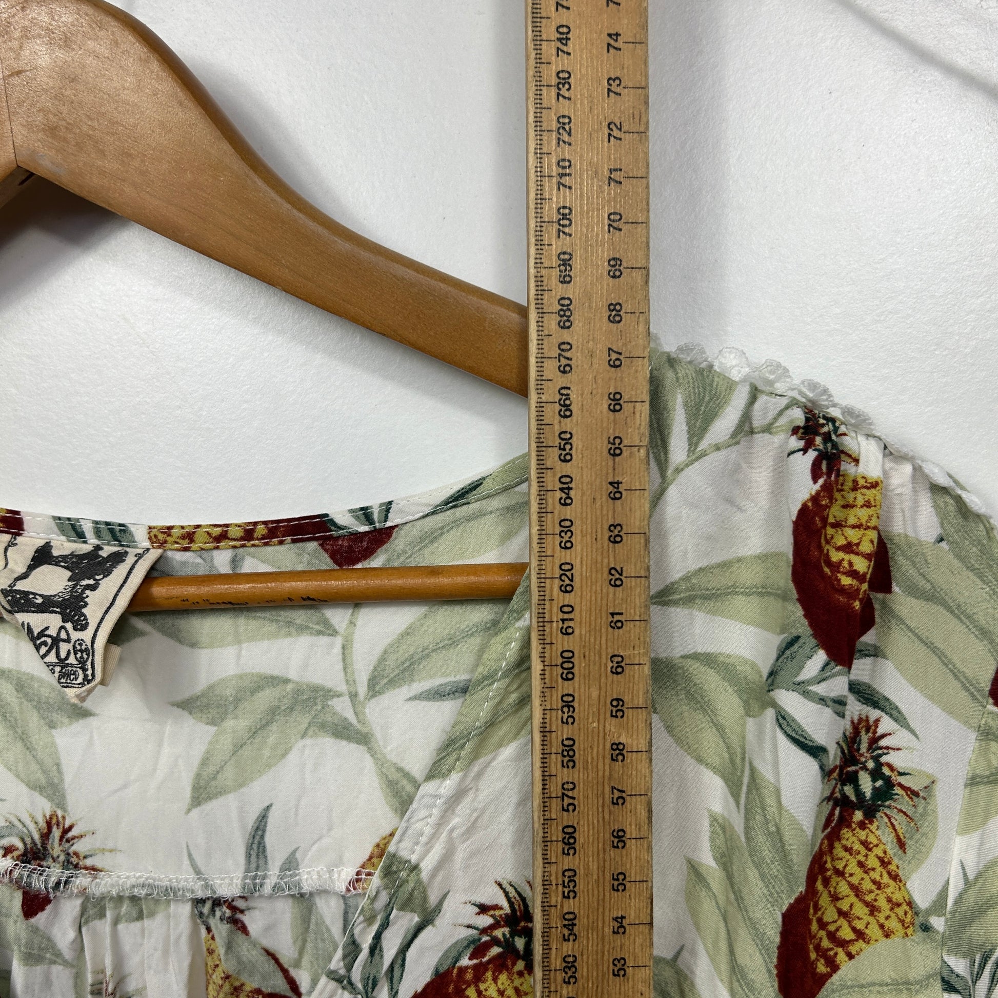 Cream pineapple and floral print top front view with wooden hanger and measuring tape